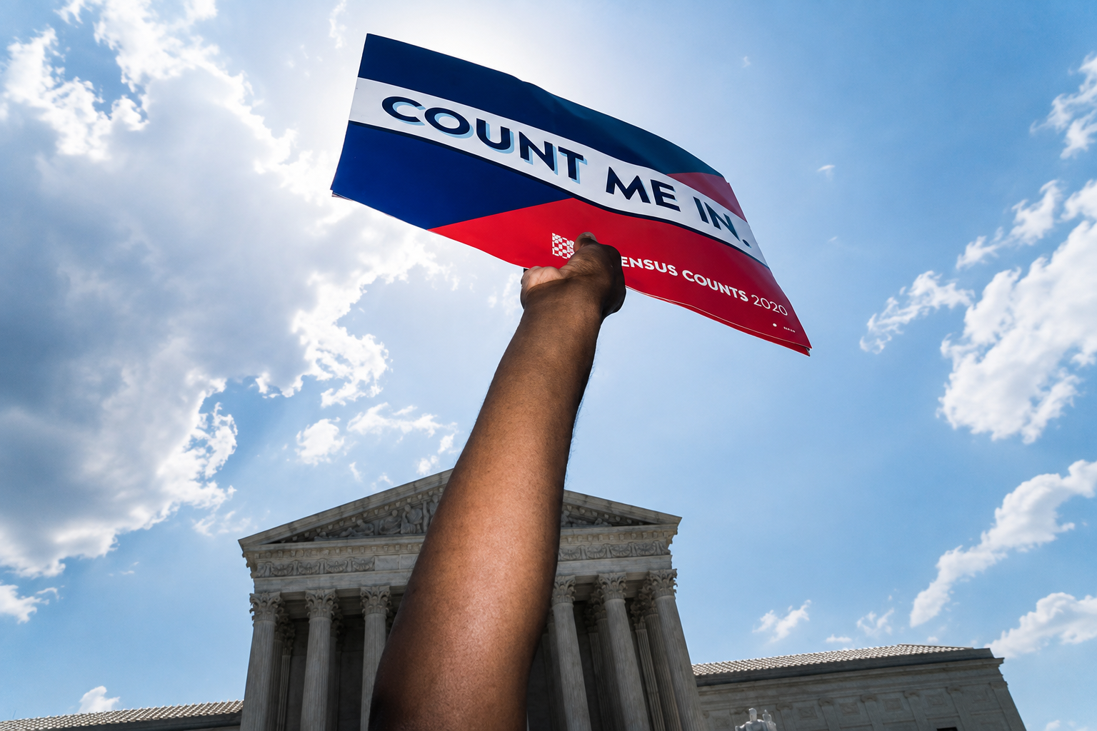 A raised hand holding a Census 2020 'Count Me In' flag in front of a federal building